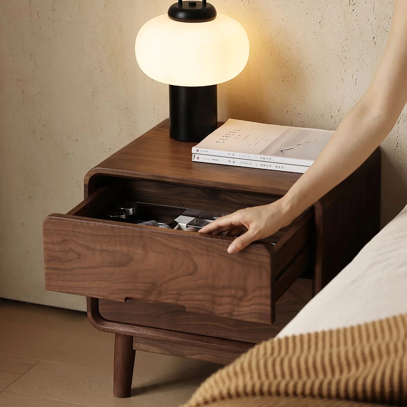 Hand opening drawer of solid walnut nightstand with lamp and books on top in cozy bedroom