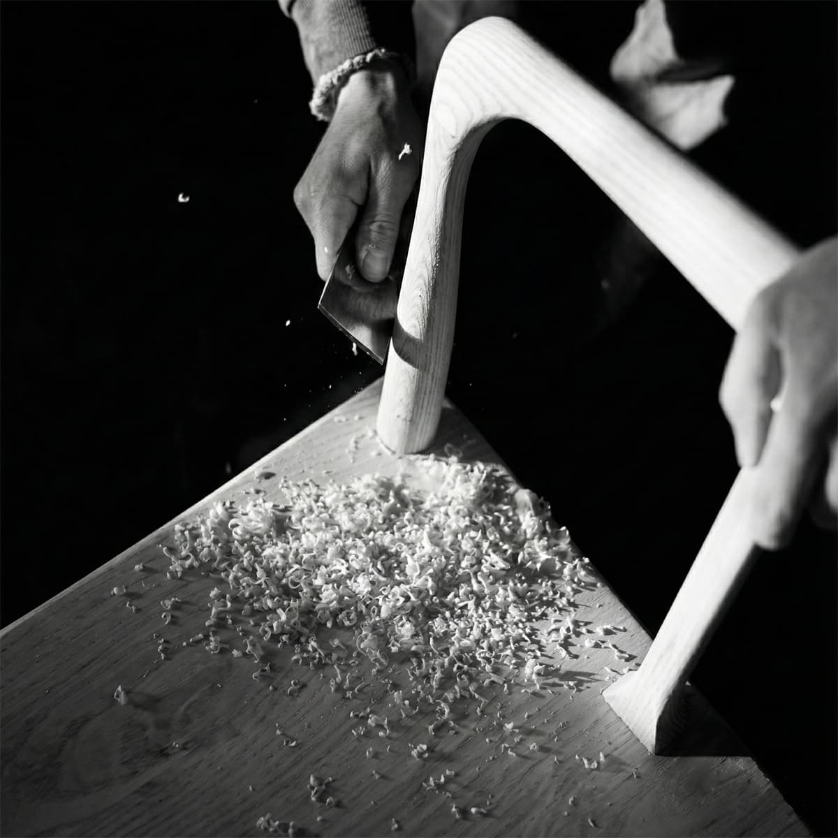 Black and white close-up of a craftsman hand-shaping a curved solid wood chair leg, highlighting artisanal woodworking quality and wood shavings.