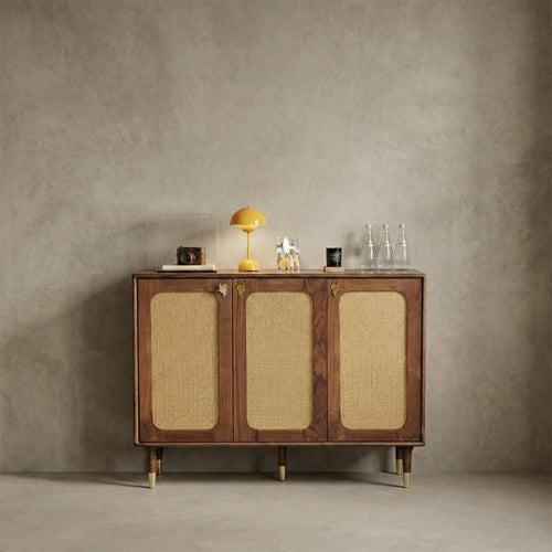 Mid-century modern walnut cabinet with woven rattan doors, brass details, yellow lamp, camera, and glass bottles