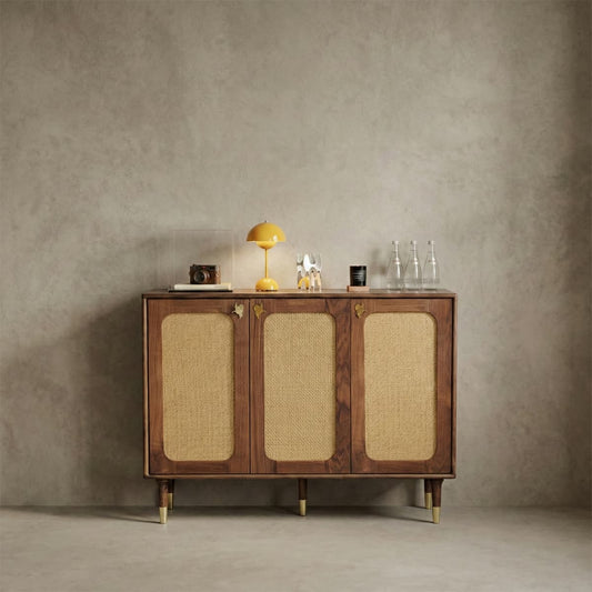 Mid-century modern walnut cabinet with woven rattan doors, brass details, yellow lamp, camera, and glass bottles