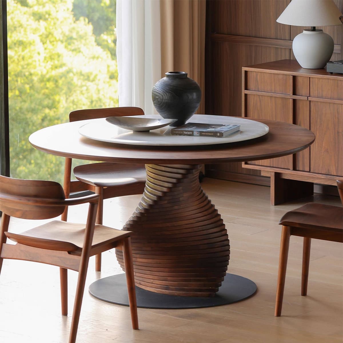 Round walnut dining table with sculpted base surrounded by mid-century modern walnut chairs in bright room