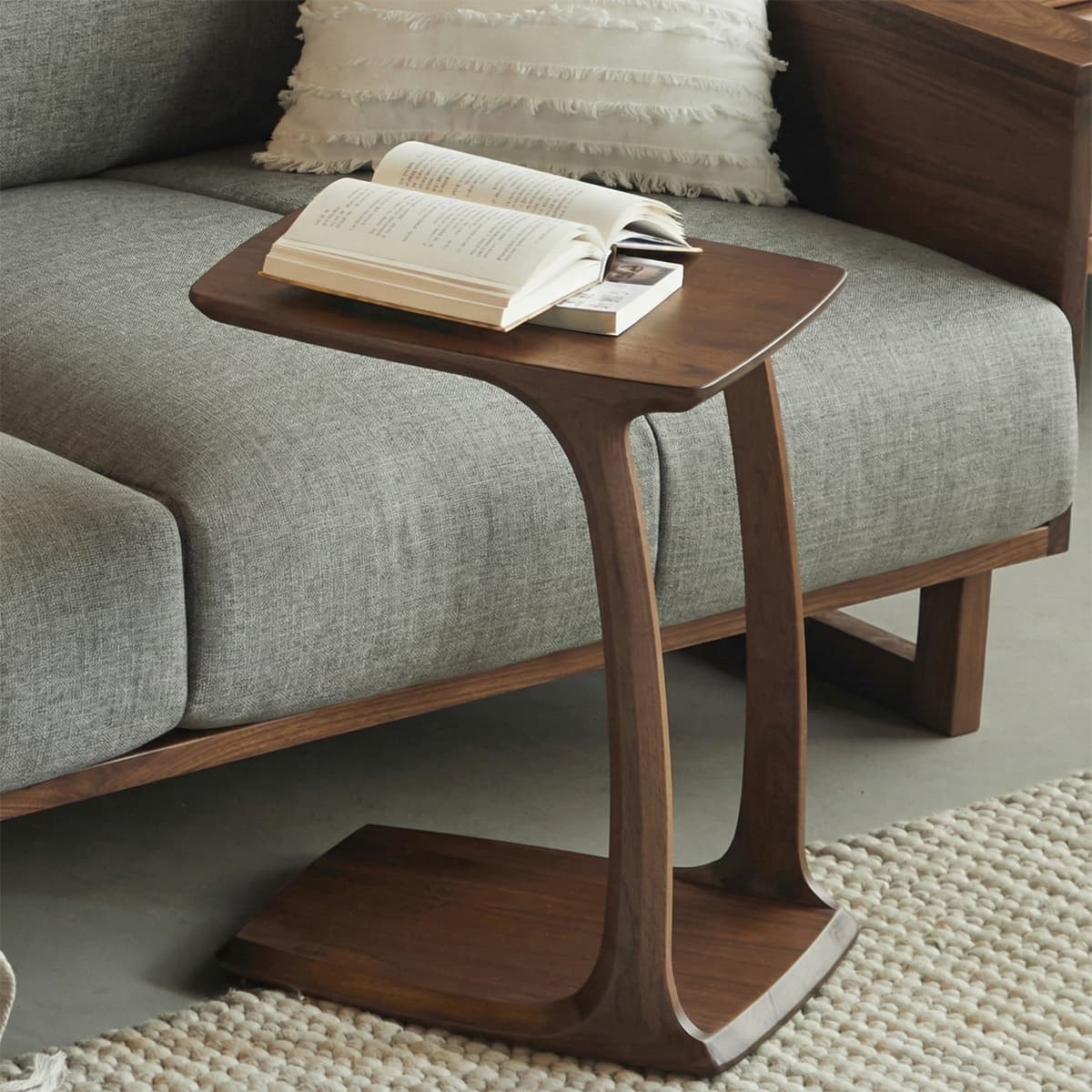 A Walnutry Helios solid North American black walnut C-table conveniently slid under a grey sofa in a modern living room, holding books.