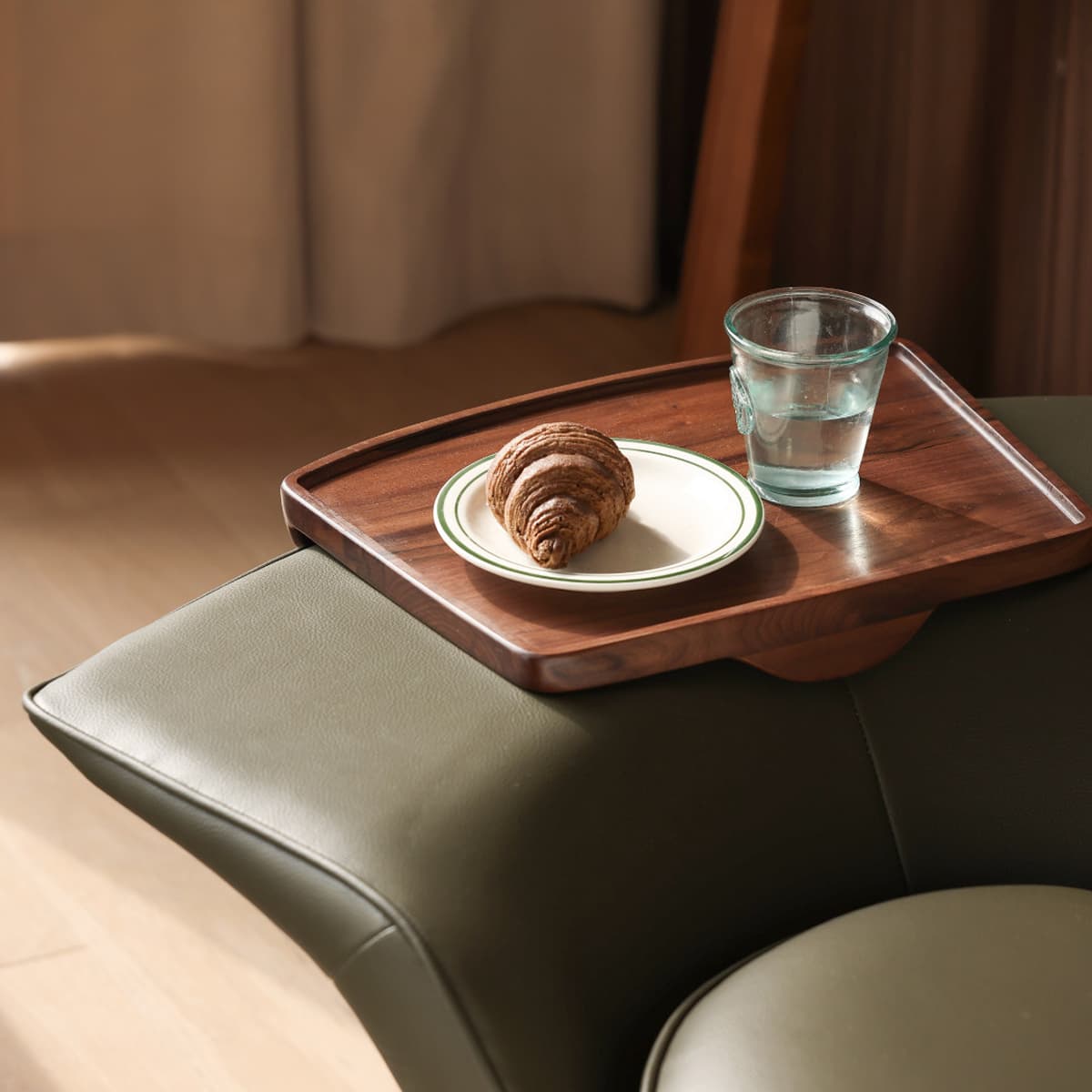 Solid black walnut wooden tray on olive green leather furniture with croissant on a plate and a glass of water