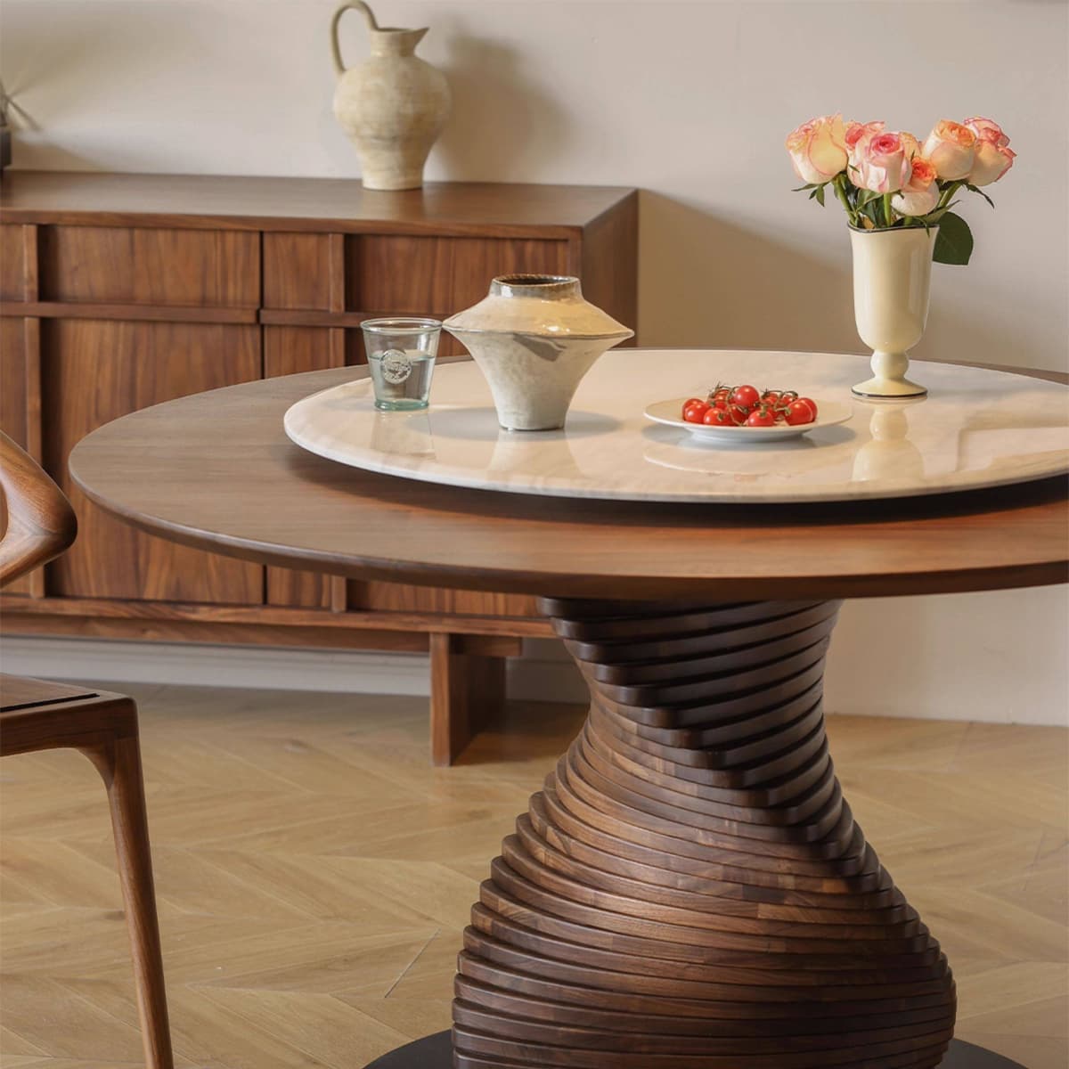 Round solid North American black walnut dining table with sculpted pedestal base, on wood floor with ceramic vase and flowers