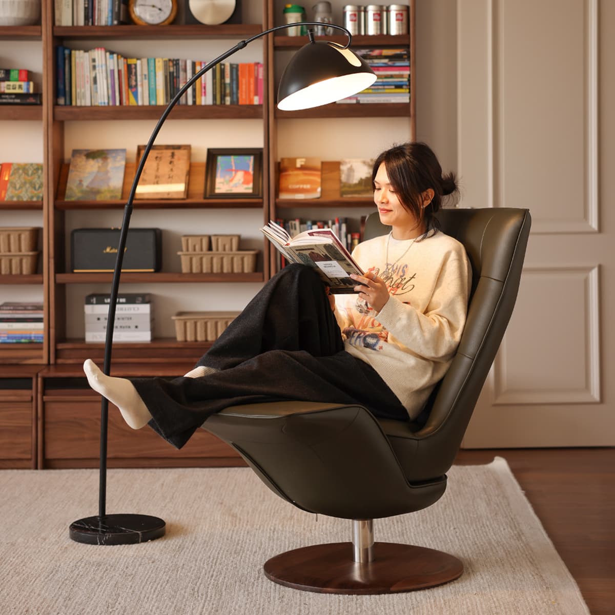 Woman reading book in modern dark leather swivel chair with floor lamp and walnut bookshelves