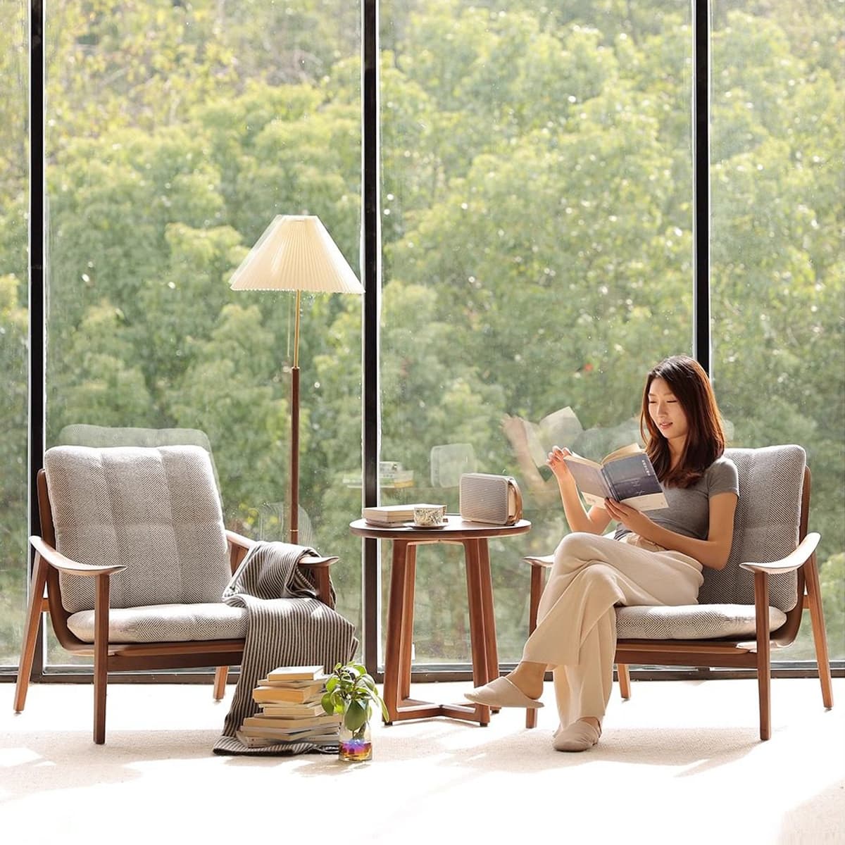Woman reading on a fabric cushioned solid walnut armchair next to matching walnut side table with lamp and books
