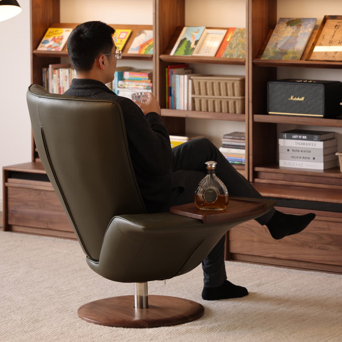 Man seated in a modern olive leather swivel chair with walnut base, holding a glass of whiskey in a cozy room with walnut bookshelves and decor