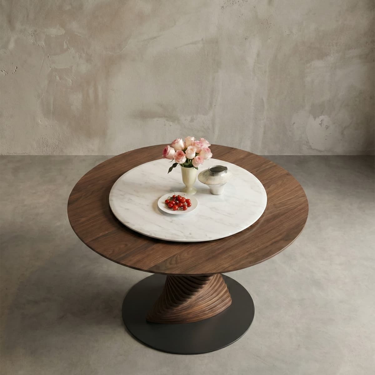 Round walnut wood dining table with a white marble lazy susan, pink roses in vase, ceramic pot, and plate of cherry tomatoes on concrete floor