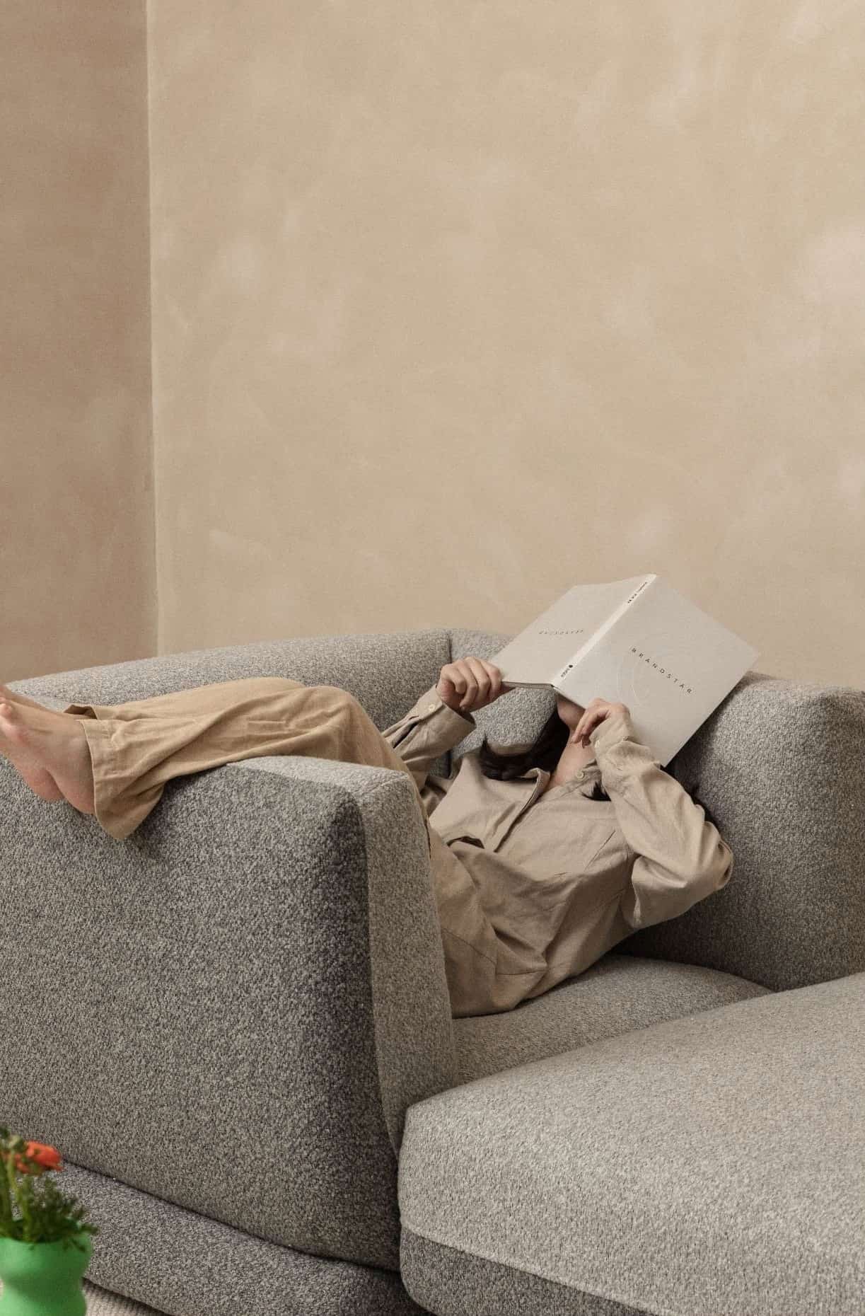 Cozy lifestyle shot of a woman relaxing on a grey fabric sofa, covering her face with an open book, wearing a beige shirt.