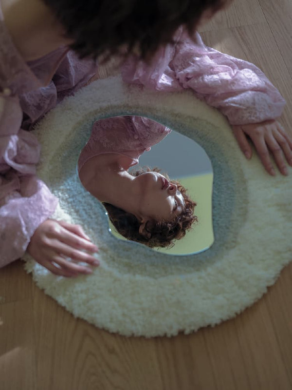 High-angle artistic shot of a curly-haired woman looking into a unique irregular-shaped floor mirror with a fluffy white and blue tufted frame.