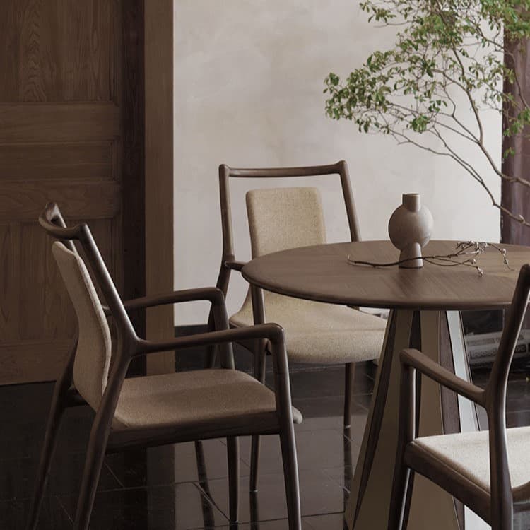 Minimalist dining room setting featuring a round walnut dining table and wooden chairs with beige fabric upholstery, styled with a ceramic vase and greenery.