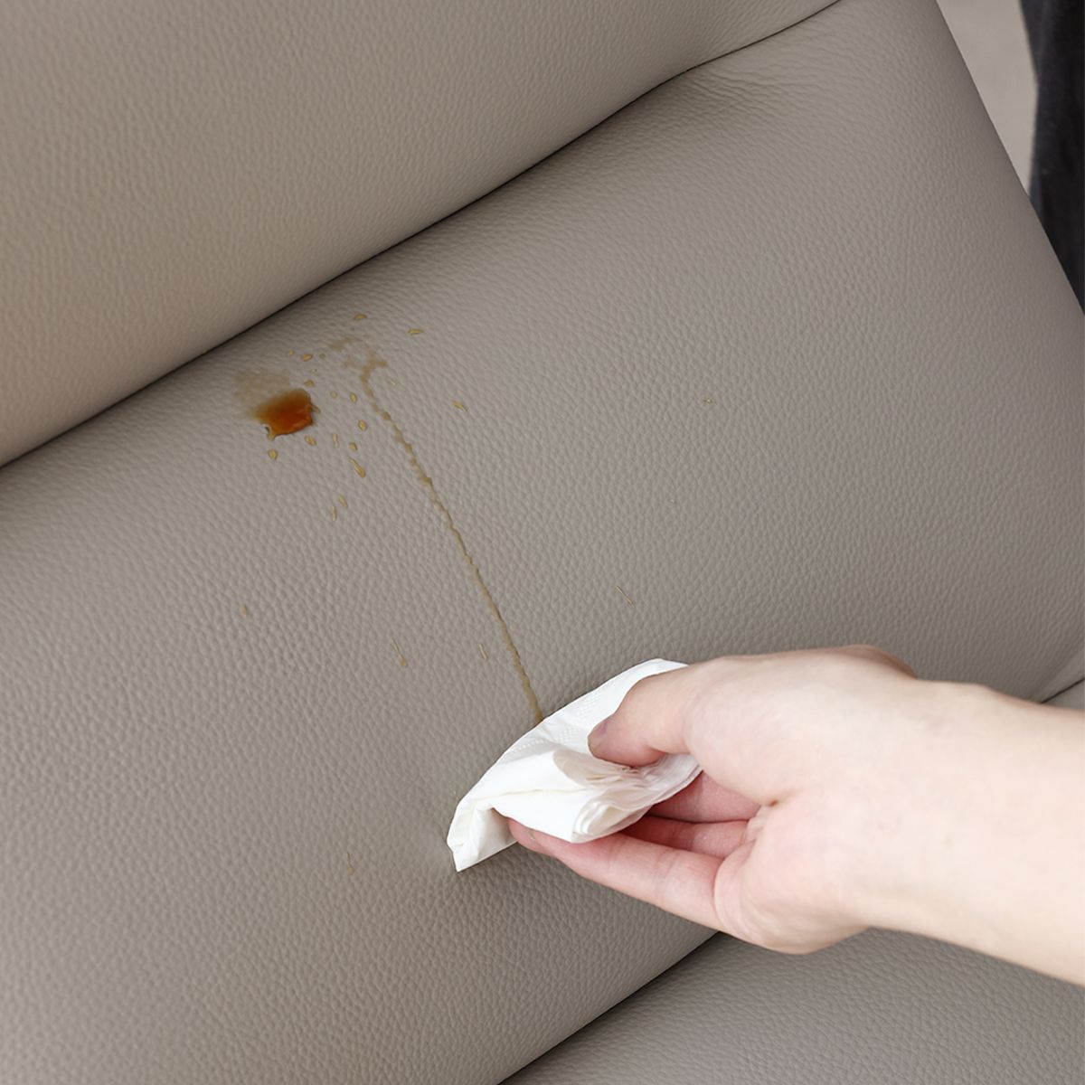 Hand wiping brown liquid spill on light grey leather surface with white cloth