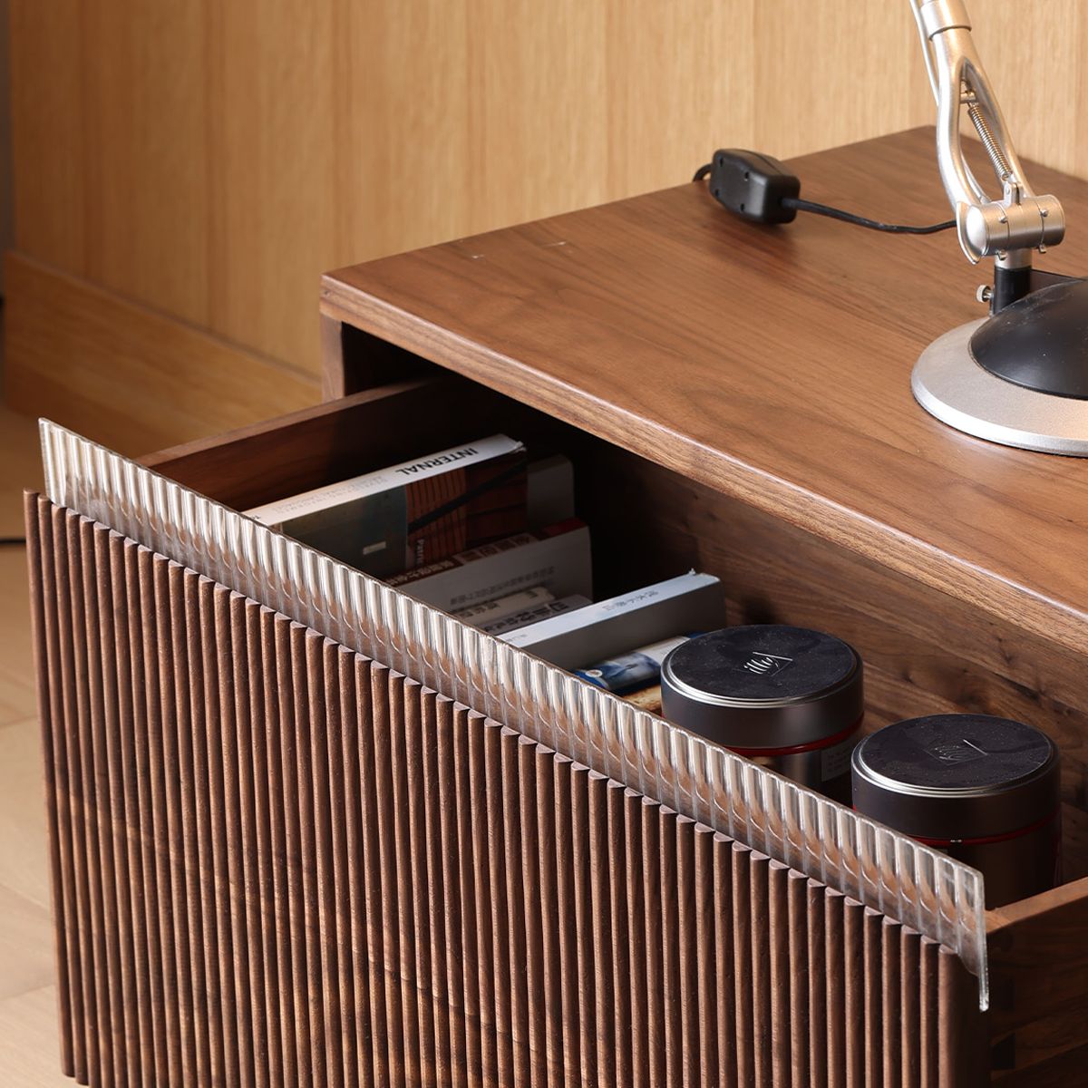 Open wooden drawer with corrugated front, holding books and two metal tins on wooden table