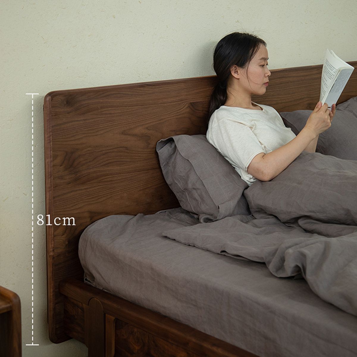 Woman reading book on bed with 81cm tall wooden headboard and gray bedding