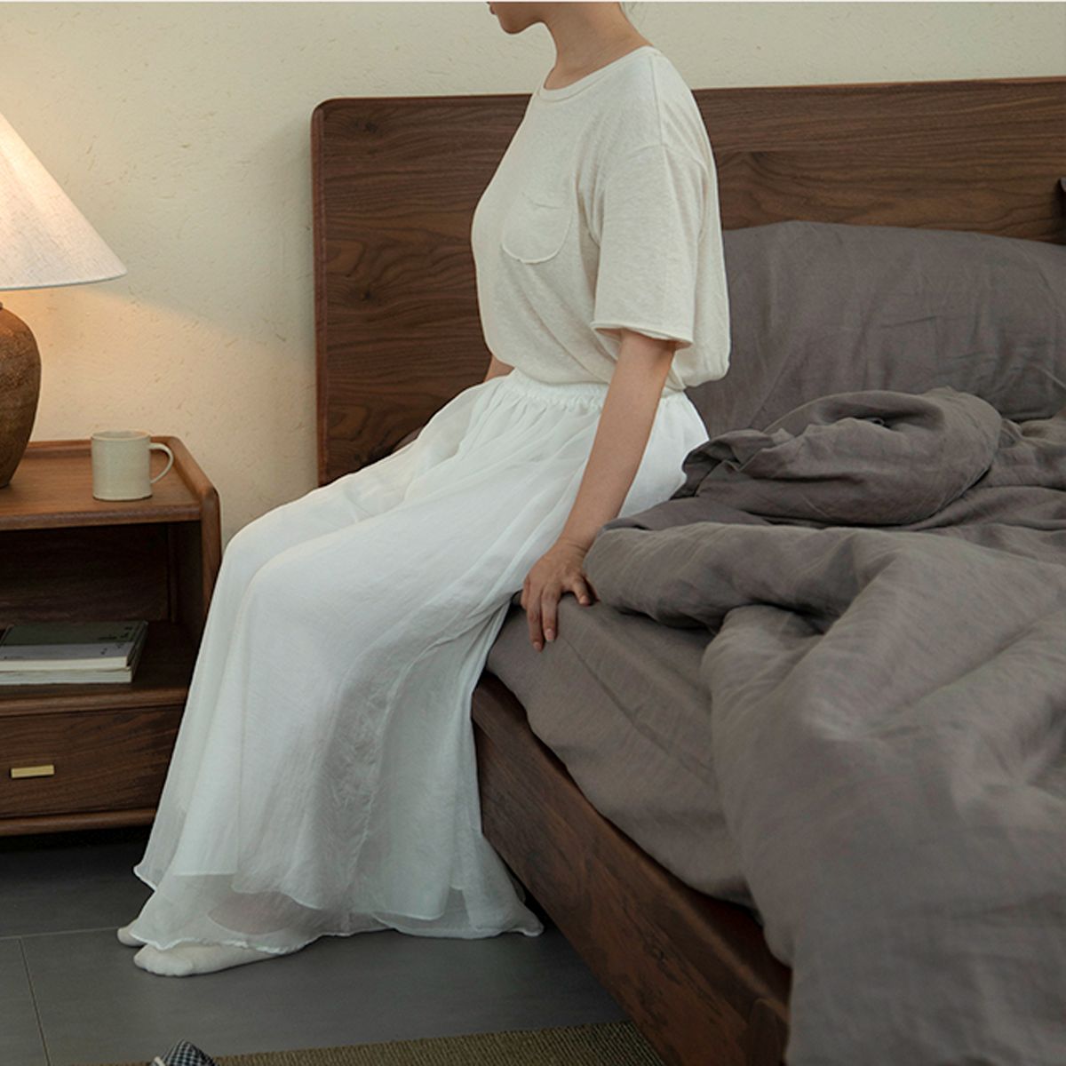 Woman in white dress sitting on edge of wooden bed with gray bedding in cozy bedroom