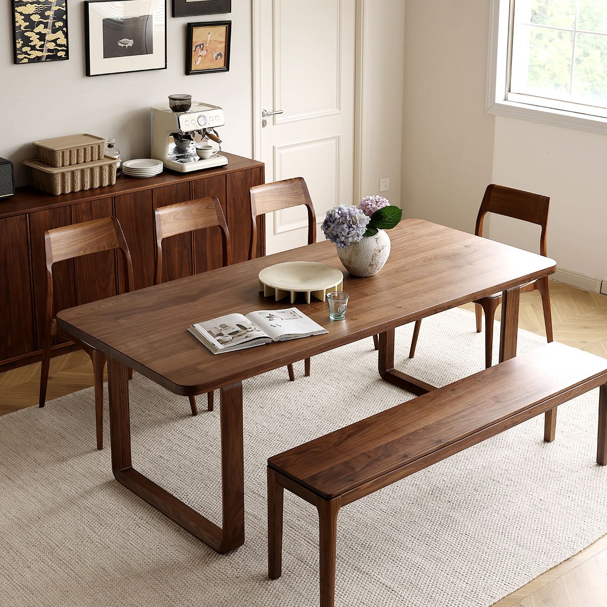Modern dining room with wooden table, bench, four chairs, vase with flowers, and coffee machine on sideboard