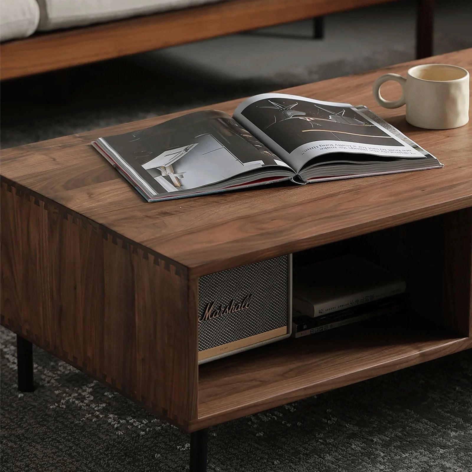 Solid black walnut coffee table with dovetail joints, open shelf holding a Marshall speaker and books, with a mug and open magazine on top
