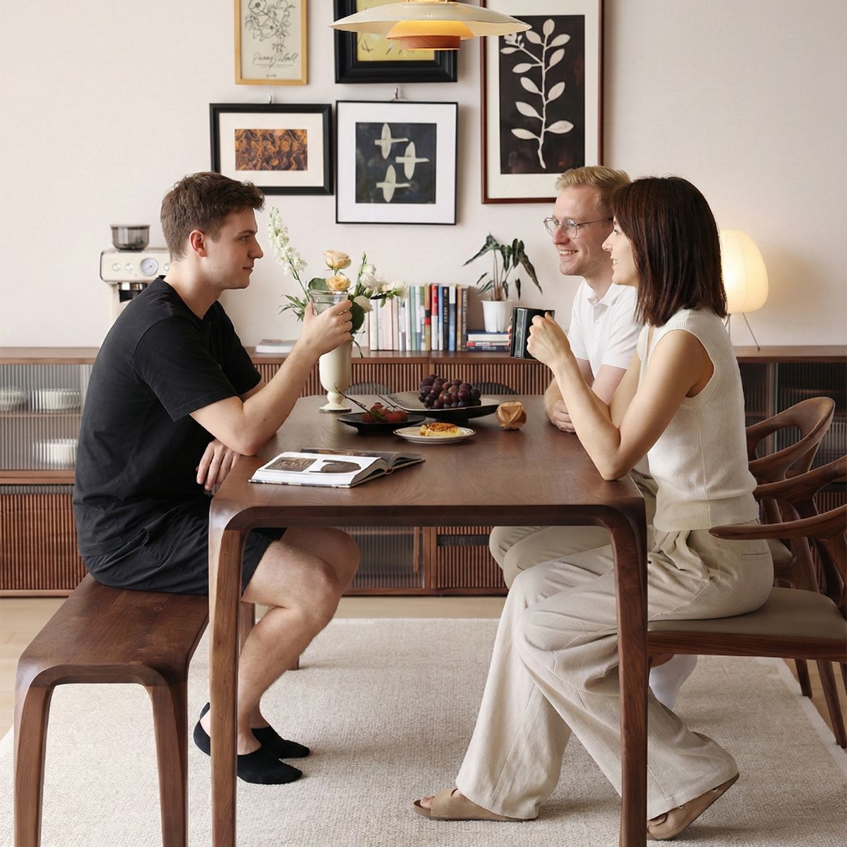 Three adults sitting around wooden dining table enjoying drinks in a modern, cozy living room with wall art and bookshelf