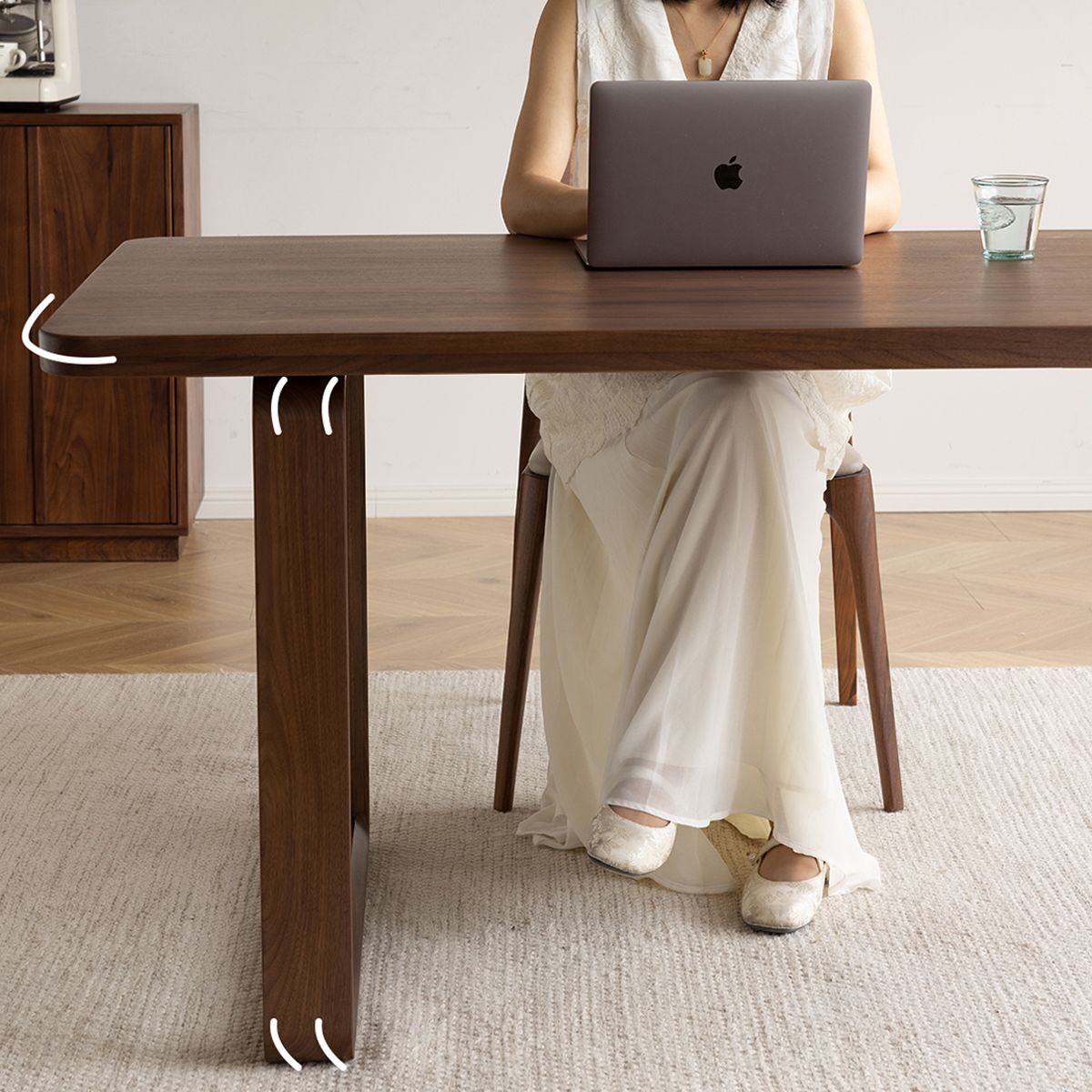 Woman in white dress using MacBook at wooden dining table with glass of water on beige rug