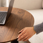 Woman's hand with gold bracelet and manicured nails resting on round wooden table near open laptop
