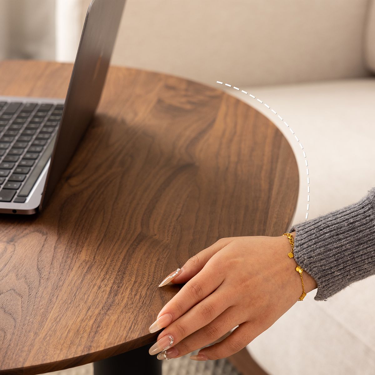 Woman's hand with gold bracelet and manicured nails resting on round wooden table near open laptop