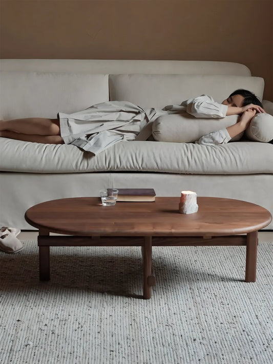 A cozy organic modern living room with a woman resting on a sofa behind the solid walnut Suiseki coffee table, illustrating the grounded living lifestyle.