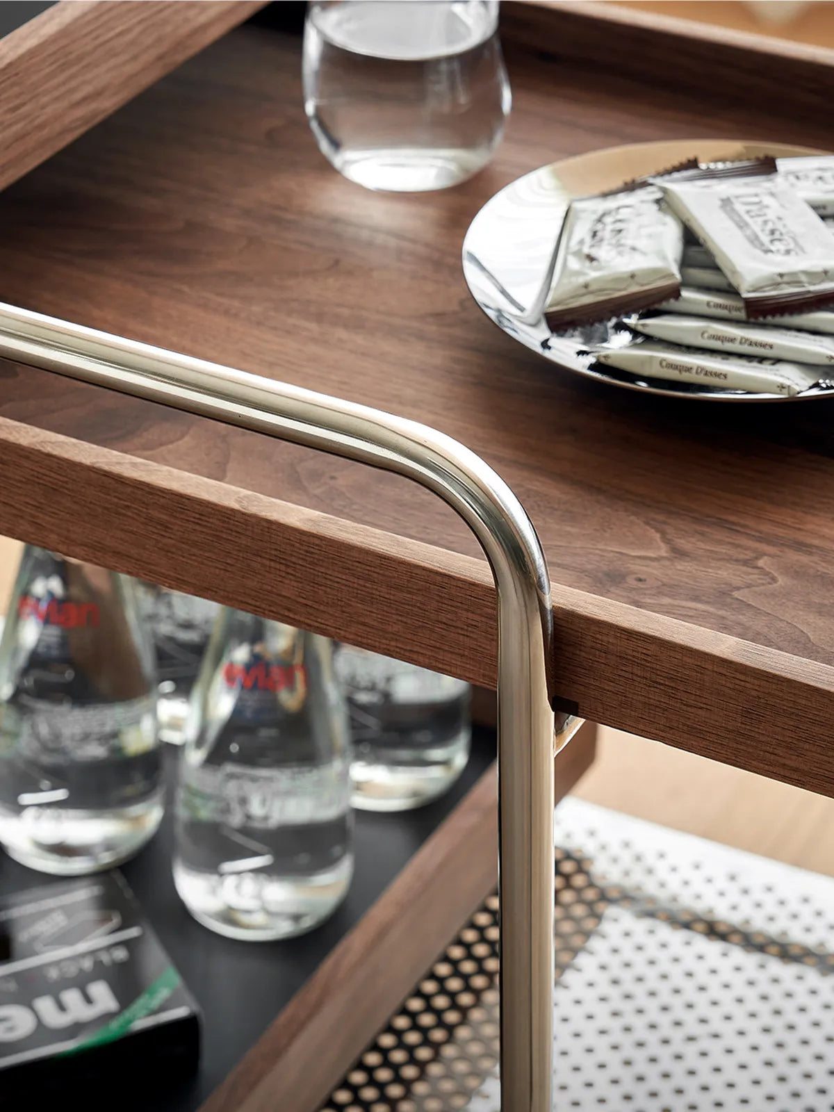 Close-up of the bottom shelf of the walnut bar cart stocked with water bottles and hospitality items.