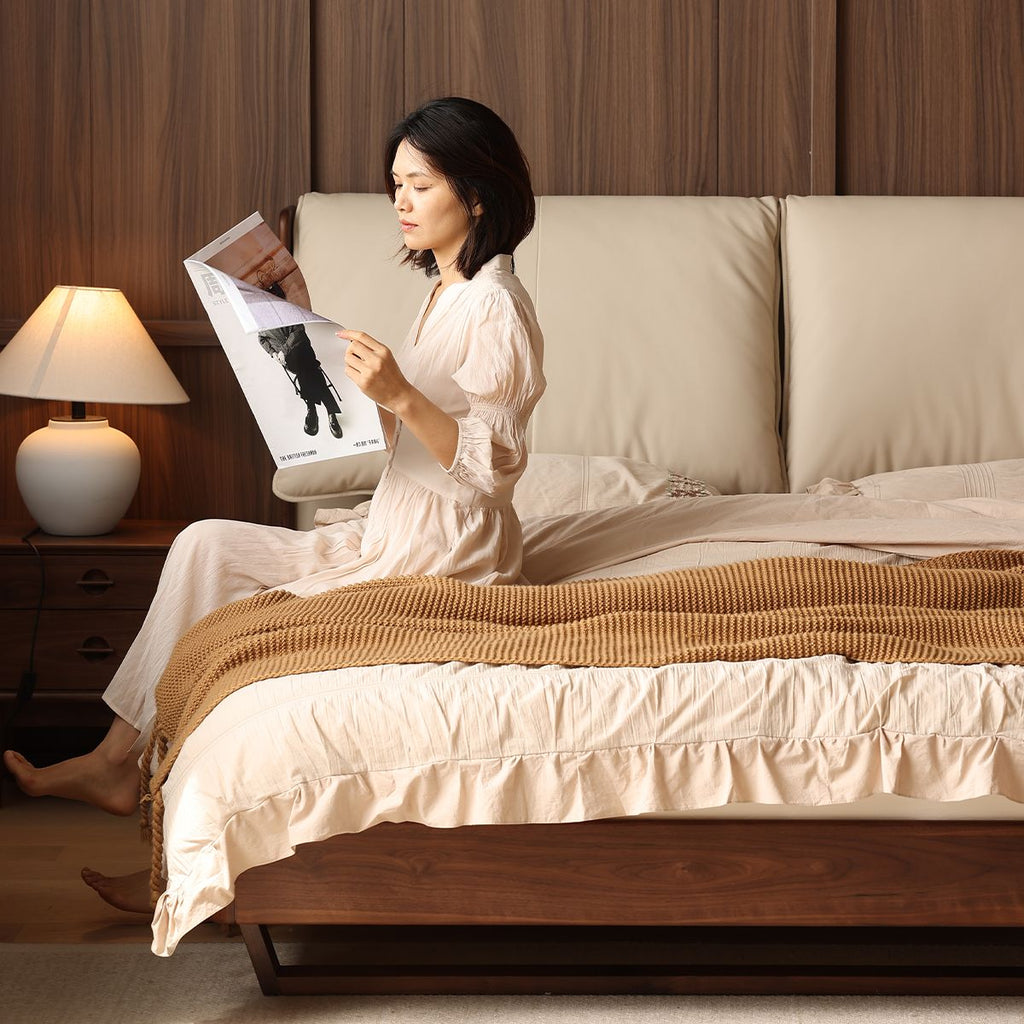 Woman in beige dress sitting on luxury wooden bed with beige bedding and reading magazine