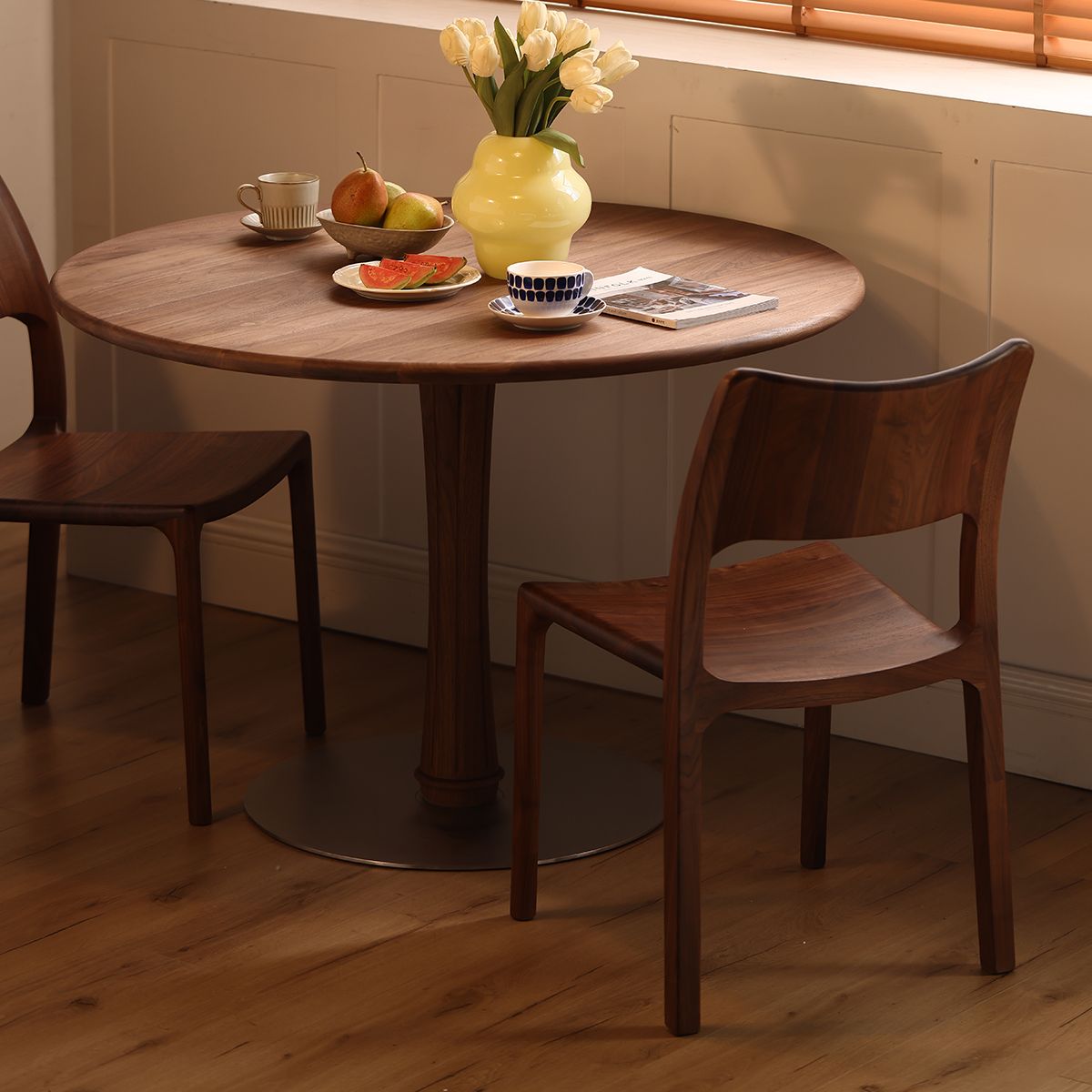 Mid-century modern round wooden dining table with two walnut chairs, yellow vase with tulips, fruit bowl, and cups