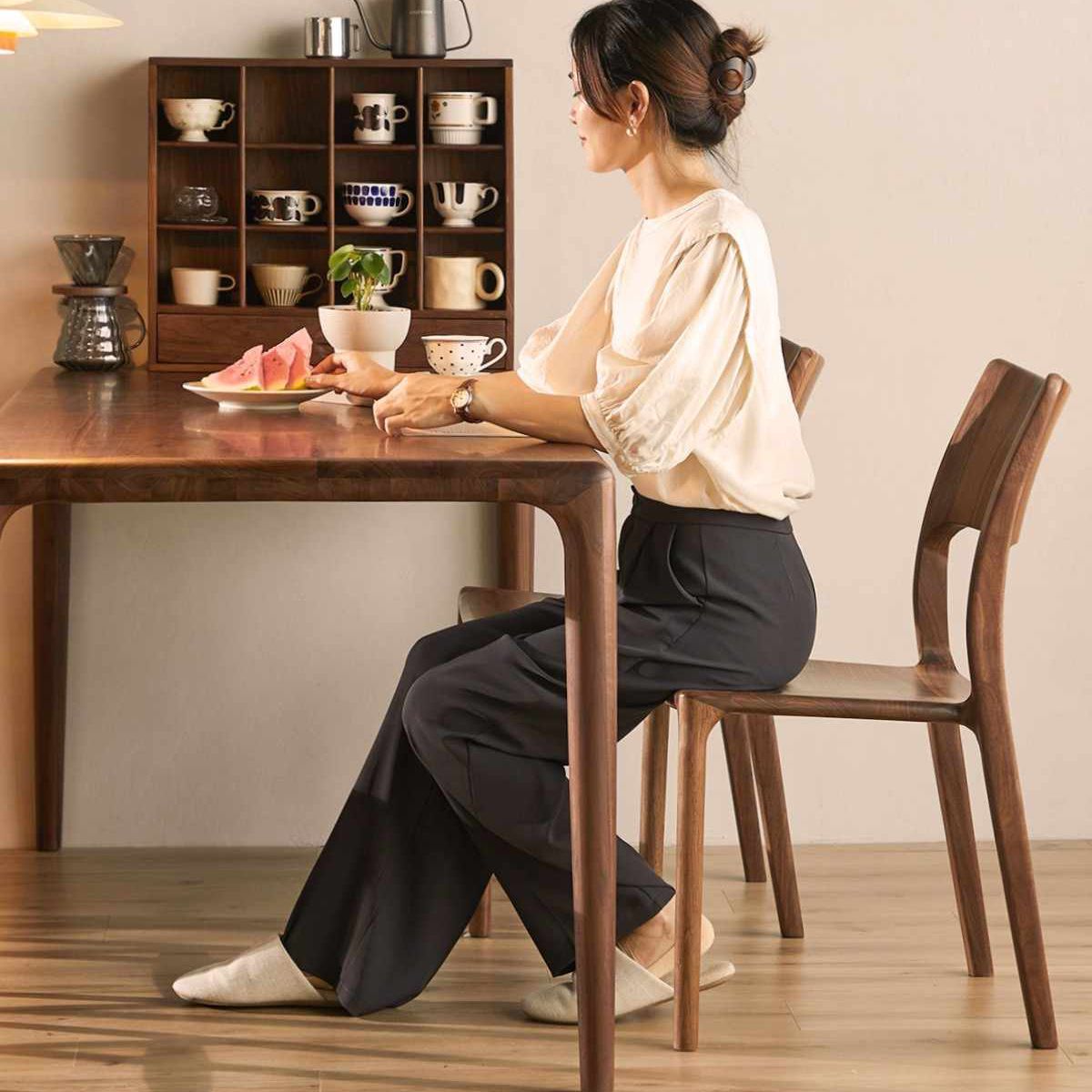 Woman seated at mid-century modern walnut dining table with matching chairs and ceramic cups display