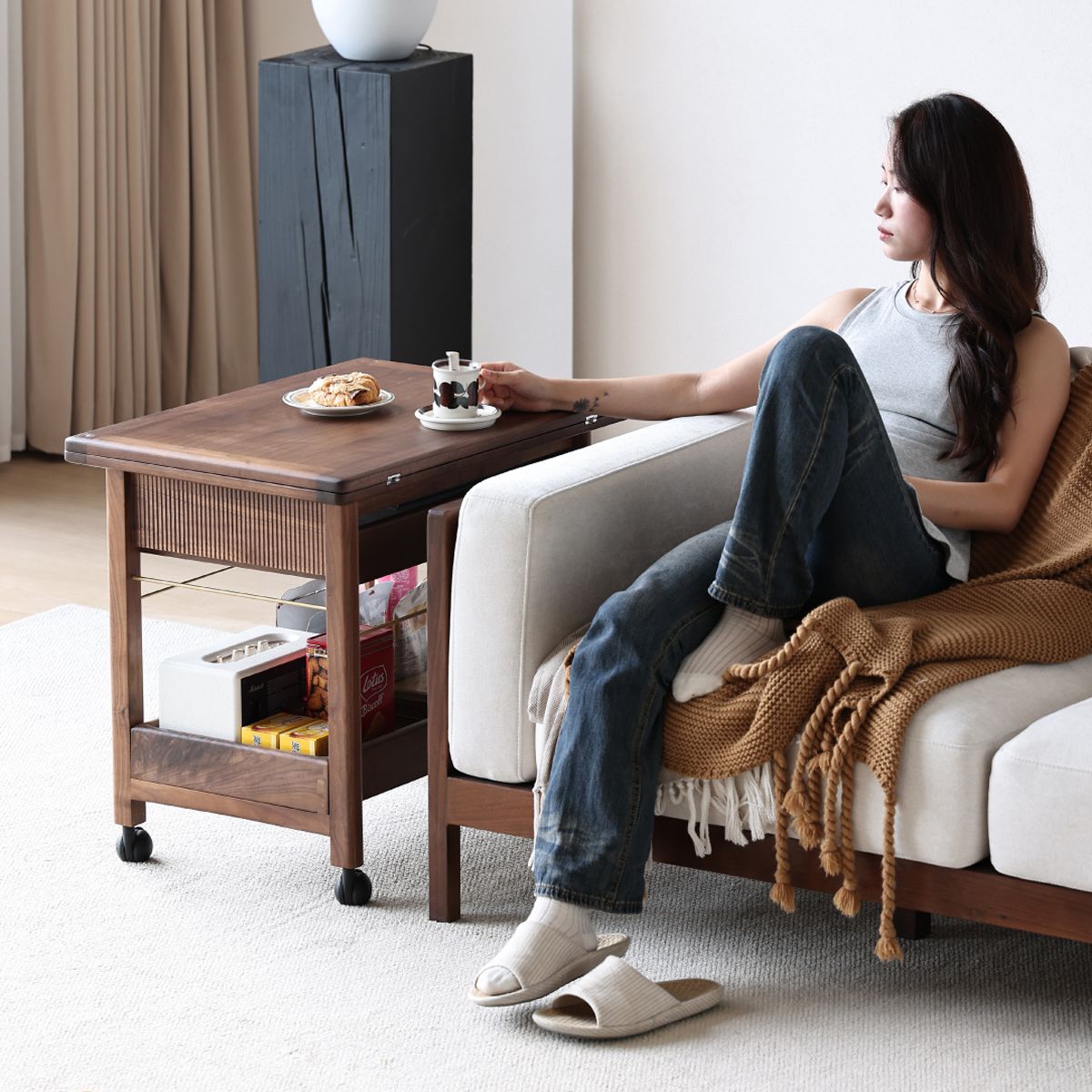 Woman relaxing on beige sofa with brown knit blanket and wooden side table holding coffee and pastry