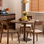 Modern dining room with round wooden table, three upholstered chairs, yellow vase with flowers, and natural light