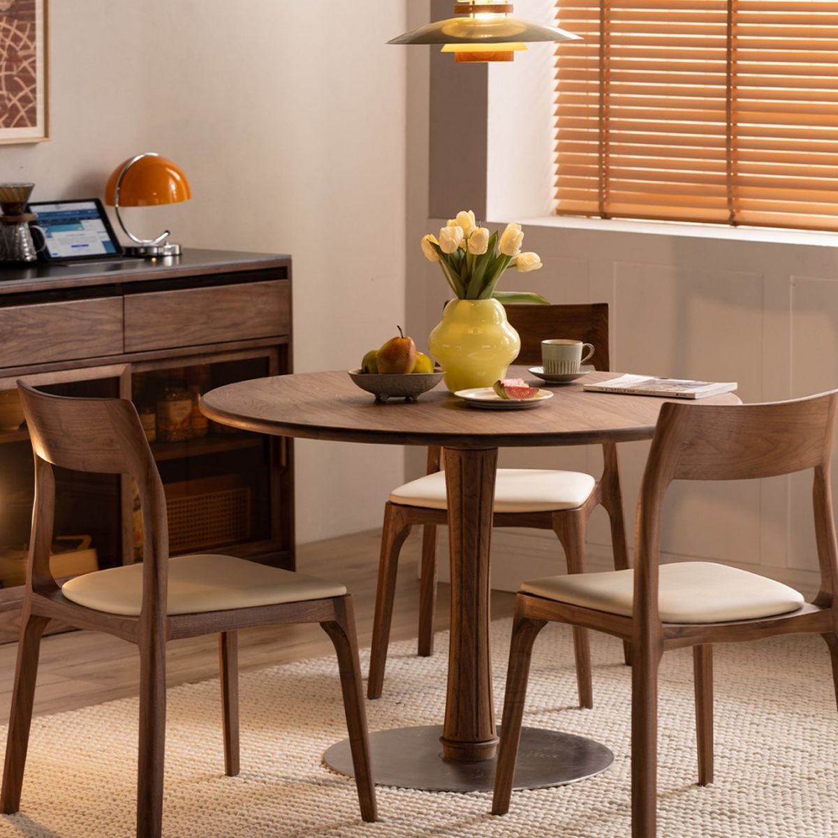 Modern dining room with round wooden table, three upholstered chairs, yellow vase with flowers, and natural light