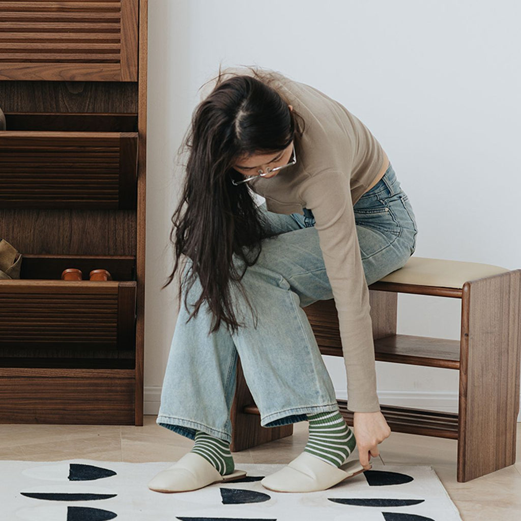 Woman sitting on wooden shoe bench putting on white slippers in modern entryway with wooden shoe cabinet