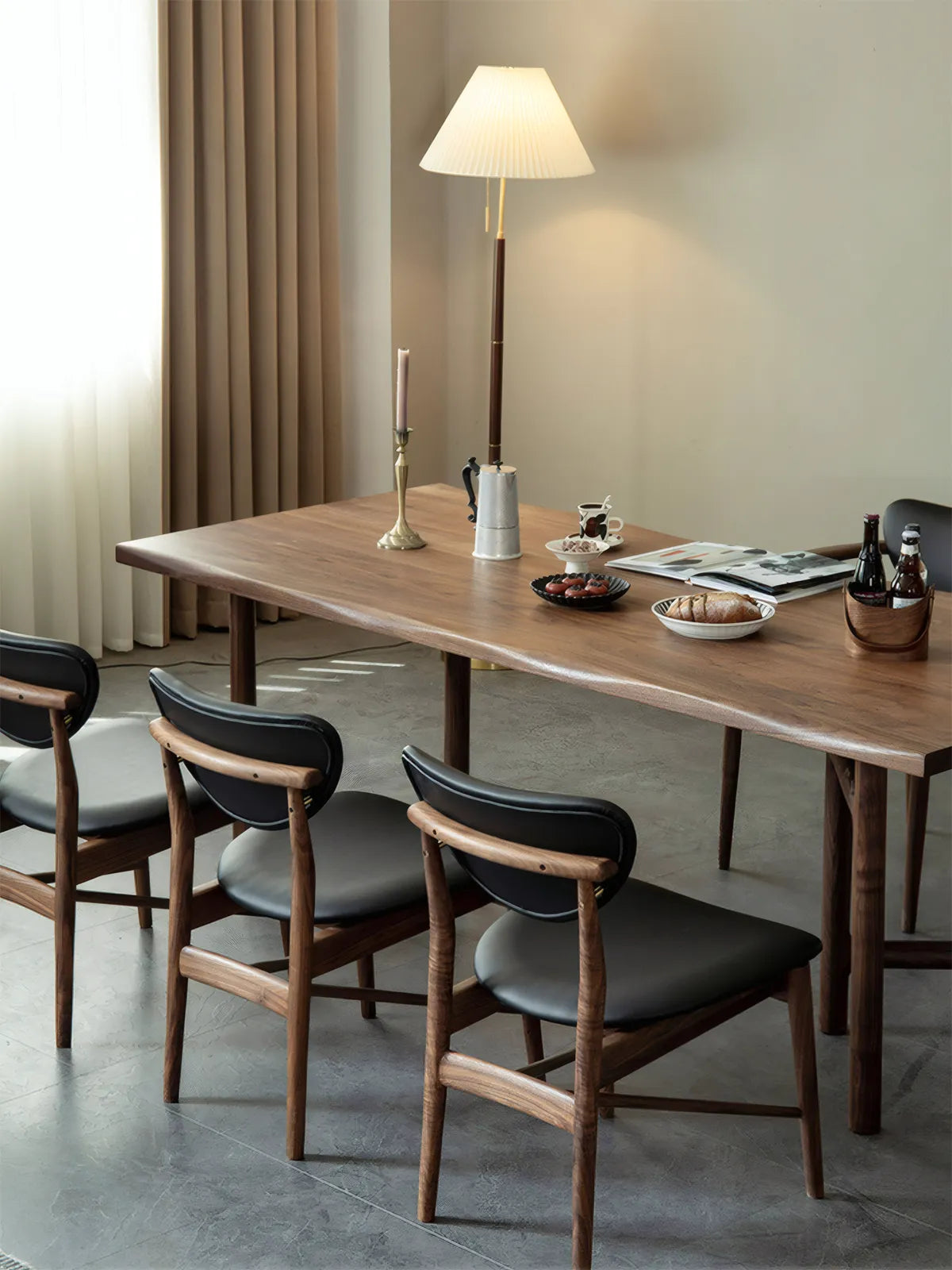 A modern dining room featuring a solid North American Black Walnut table paired with Sylvan walnut dining chairs, illuminated by a warm floor lamp.