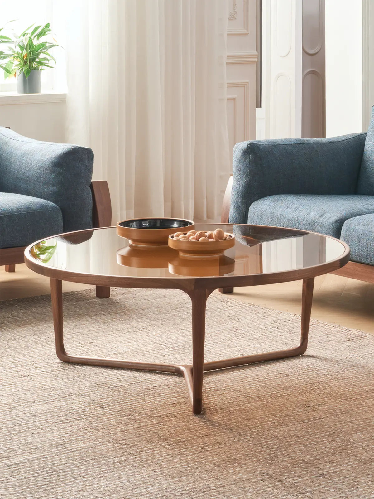 Studio isolation shot of the large round amber glass coffee table, highlighting the transparency of the top and the sculptural silhouette of the solid walnut tripod base.

