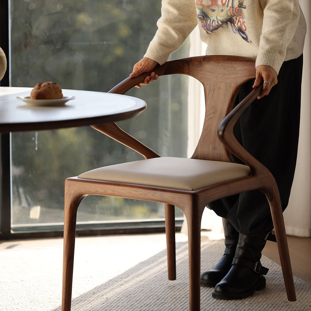 Person positioning a sculptural wooden chair with beige cushion near round table by window