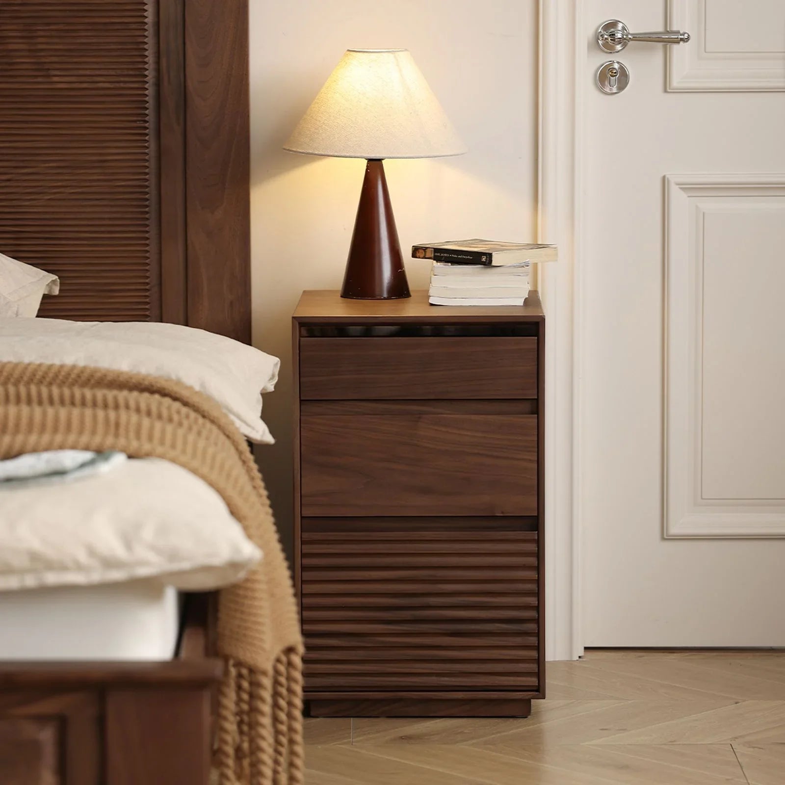 Walnut wood bedside table with lamp and books next to bed in cozy bedroom