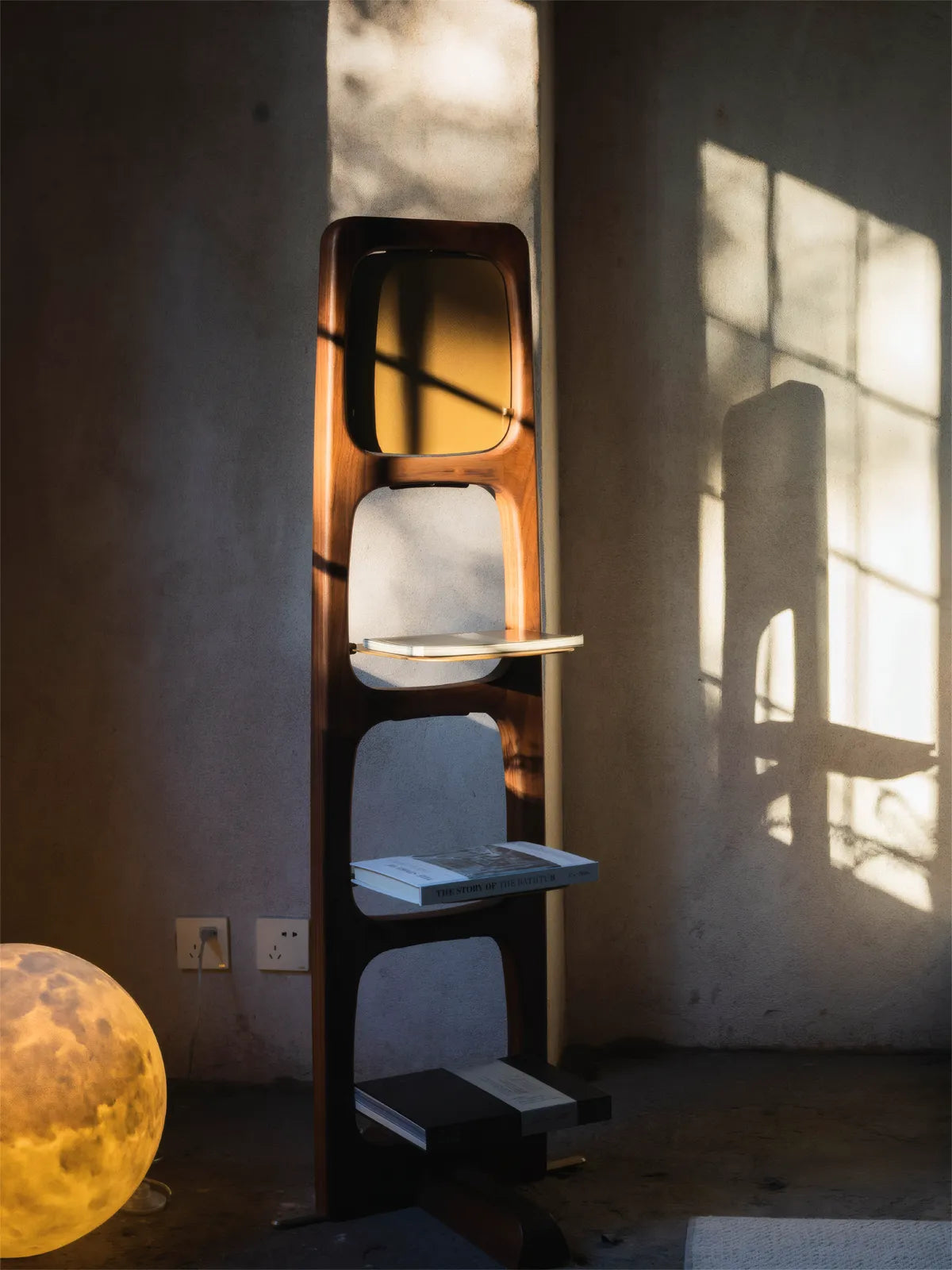Front view of the Ocular solid black walnut etagere standing in a concrete room with dramatic sunlight, highlighting the organic rounded cutouts.