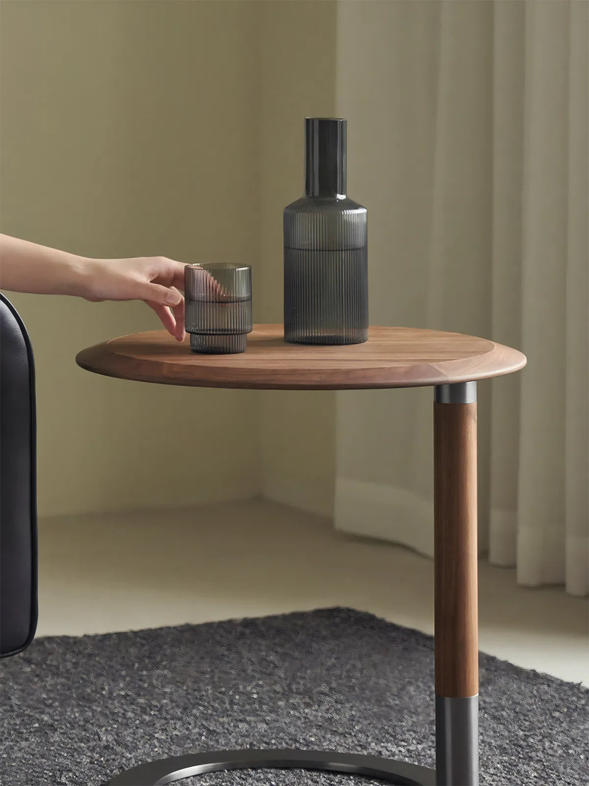 Close-up of the Suiseki round table top showing the rich grain of North American Black Walnut and a hand reaching for a ribbed glass carafe.