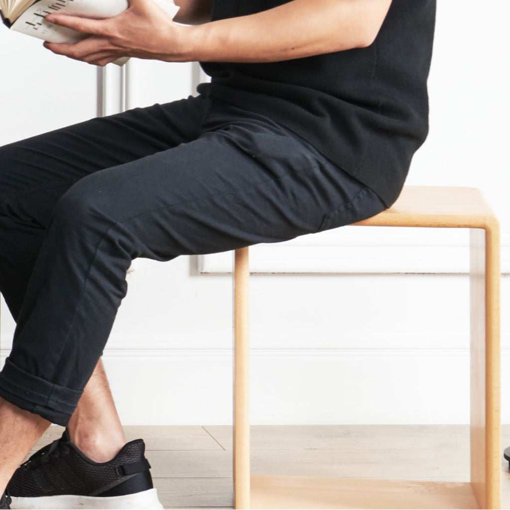 Person sitting on minimalist light wood bench wearing black pants and black sneakers reading a book