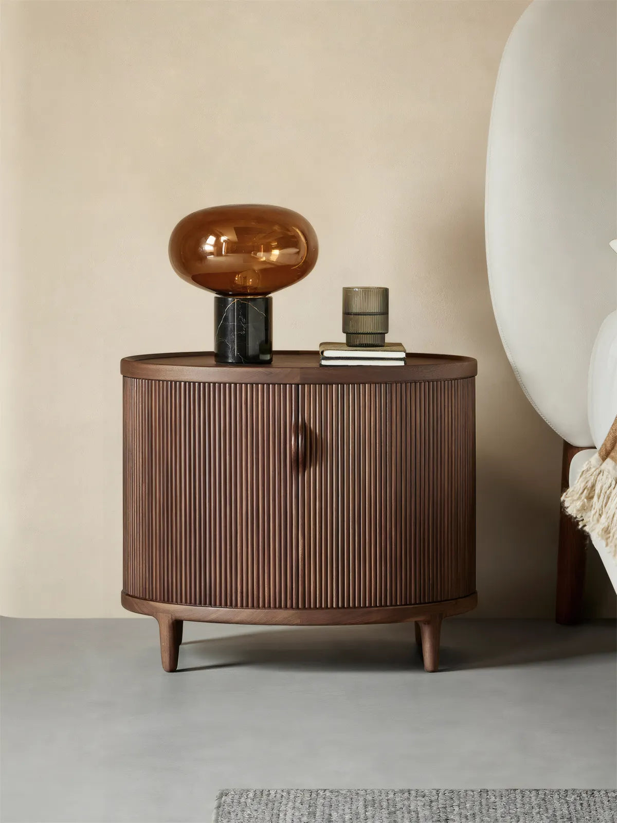 Suiseki Oval Nightstand placed next to a white bed, showcasing its mid-century modern design and solid walnut finish in a warm bedroom setting.