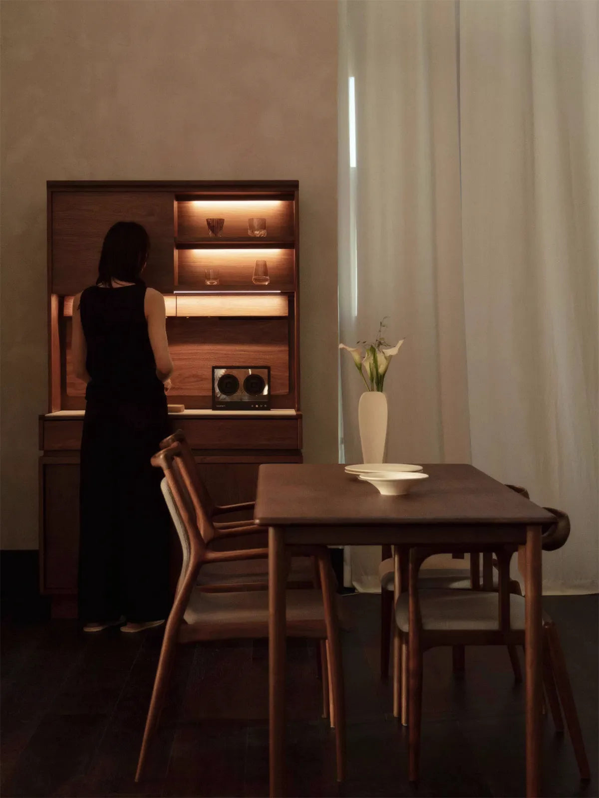 Detail of Walnutry solid walnut cabinet showing the rich matte wood grain finish and warm LED lighting, with a woman standing beside it for scale.