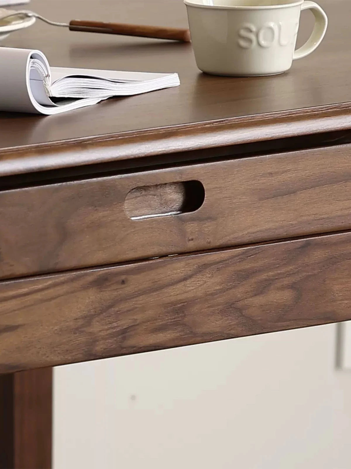 Close-up of the apron and recessed pull on a walnut extendable dining table, with a rolled magazine, a mug, and a small utensil on the tabletop.