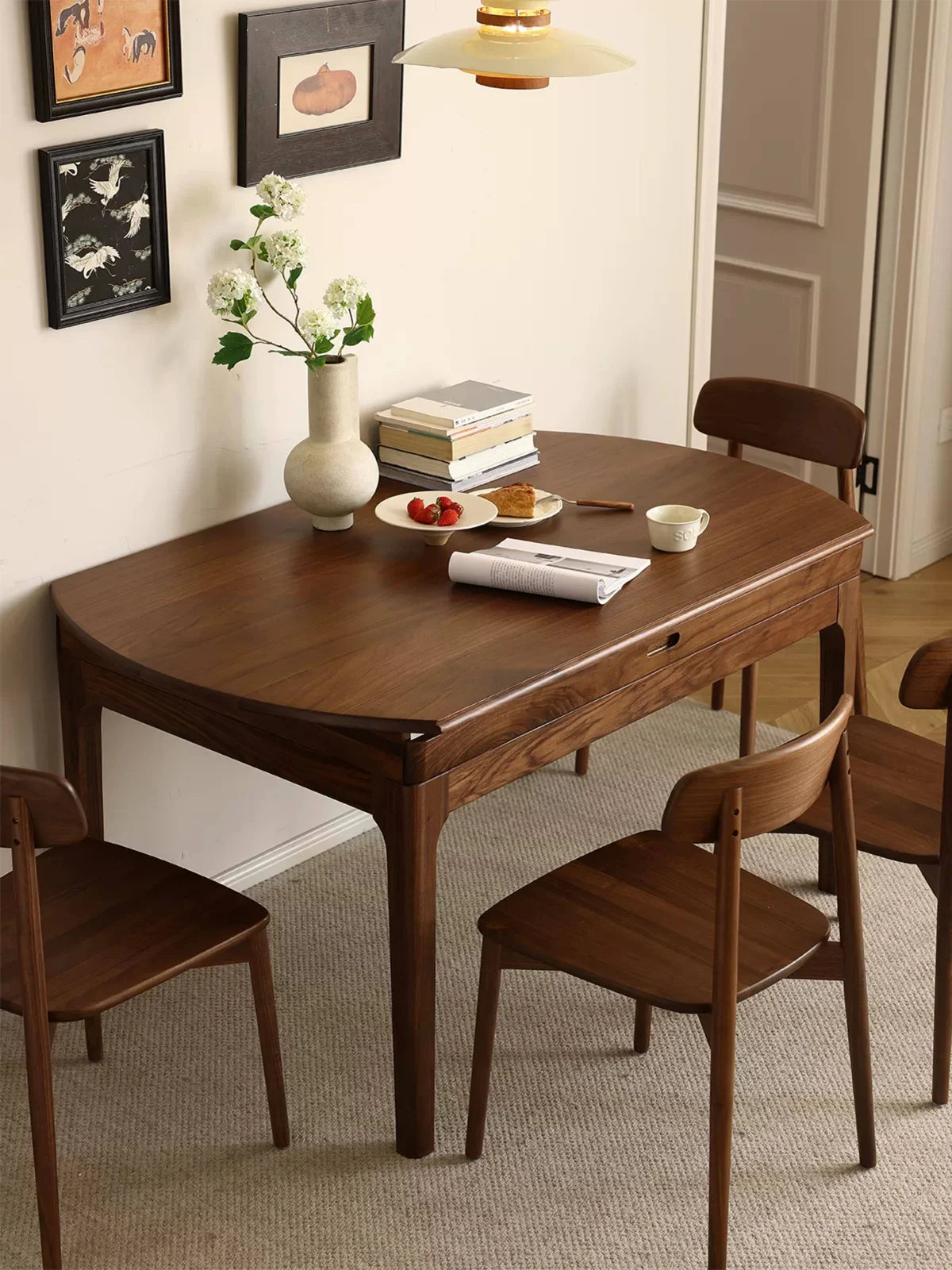 Walnut extendable dining table styled in a compact dining nook with four matching chairs, framed wall art, flowers, books, and breakfast items on top.