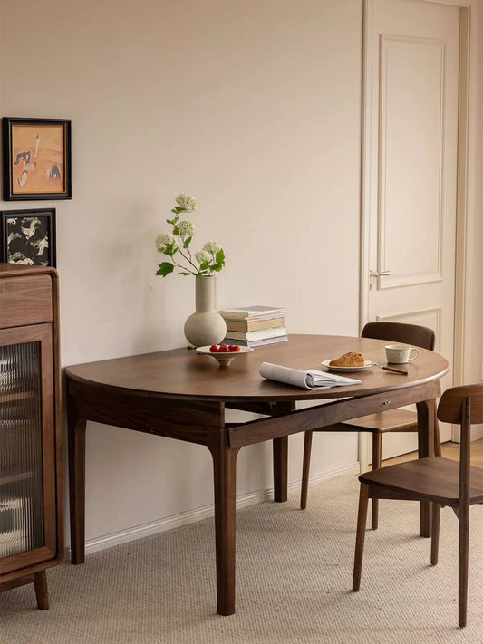 Walnut extendable dining table in a space-saving wall-side configuration, styled with flowers, books, fruit, and a pastry, with matching wooden chairs nearby.