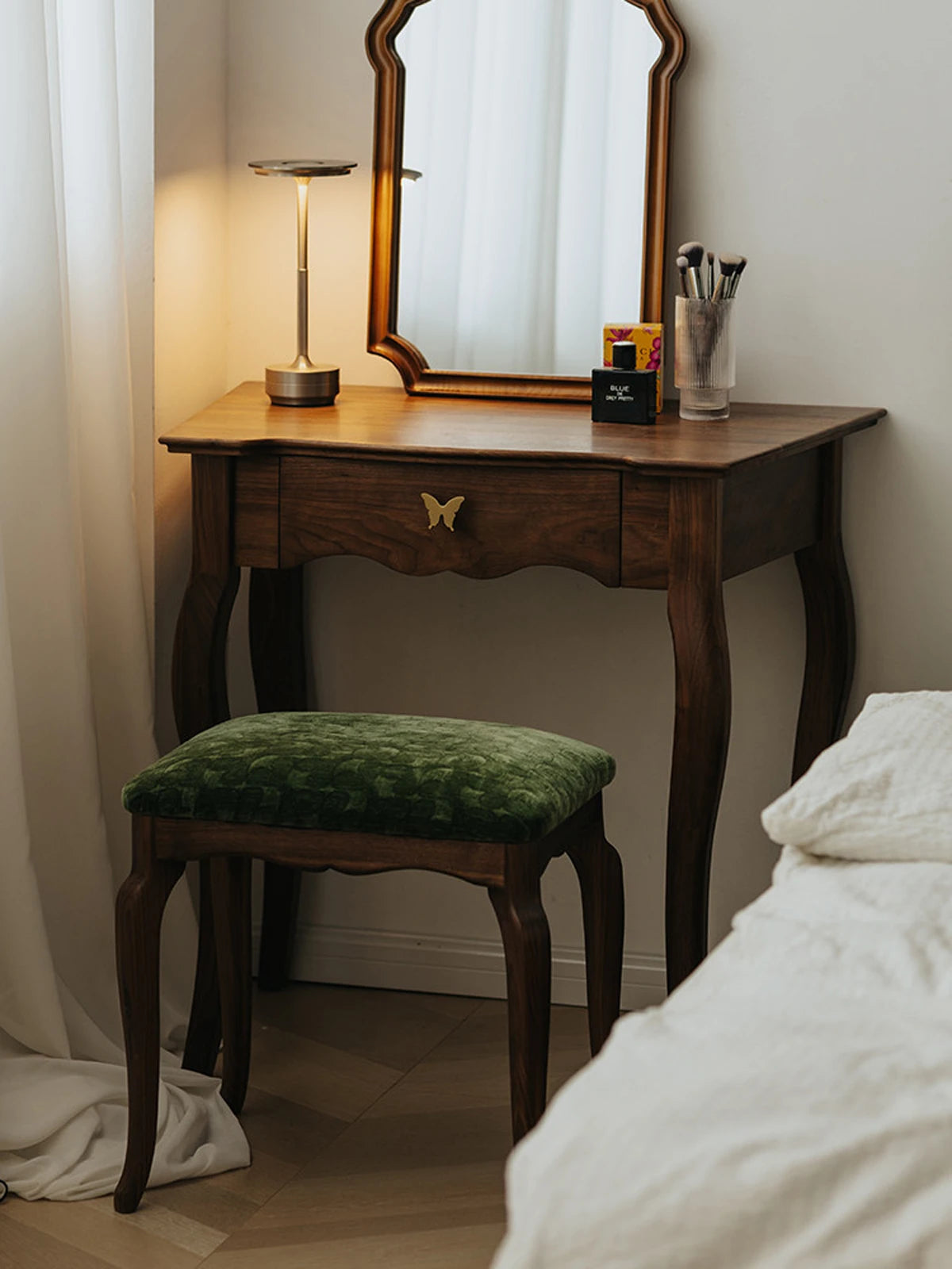 The Bourbon Vanity Stool styled in a bedroom setting, tucked under a matching wooden vanity desk with a mirror and lamp.
