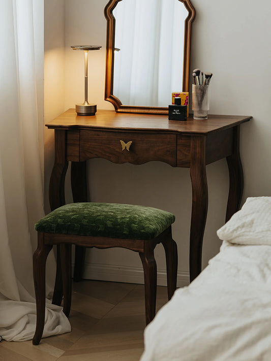 The Bourbon Vanity Stool styled in a bedroom setting, tucked under a matching wooden vanity desk with a mirror and lamp.