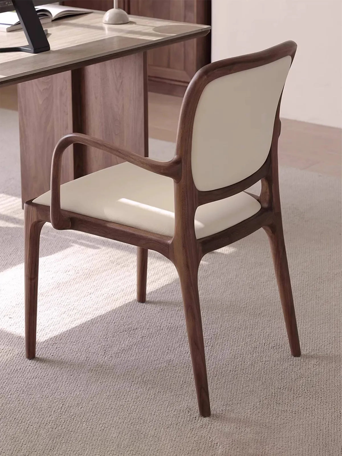Rear three-quarter view of the Cael dining arm chair beside a desk, highlighting the continuous curved walnut back frame.