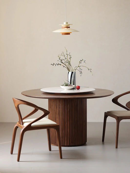 Front room view of a walnut round pedestal dining table with lazy Susan, floral vase, and pendant light above.